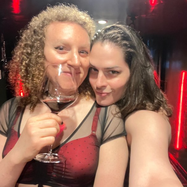 Avery (right) and Violet (left) grin at the camera while in their dungeon as Vi sips a red beverage from a stemmed wine glass. (It's probbably wine.)